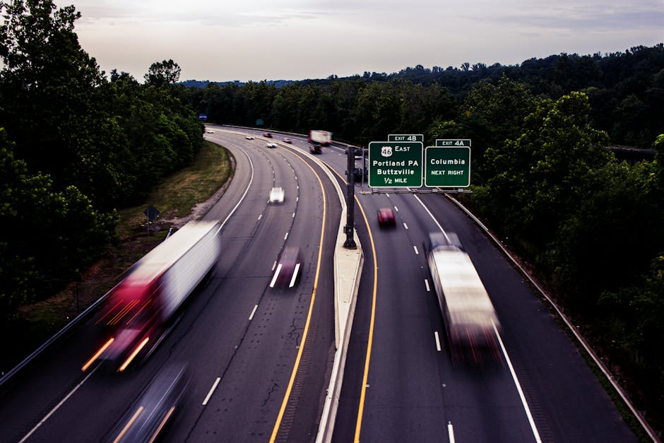 Aerial view of a multi-lane motorway with several vehicles, including a large white truck and smaller cars, traveling in both directions. Green overhead road signs indicate exits for Portland PA and Buttzville via exit 4B, and Columbia via exit 4A, with the signs positioned above the lanes. The road is bordered by dense trees on both sides, and the sky above is overcast with a mix of clouds. The scene captures the movement and flow of traffic during daylight hours, illustrating typical conditions for vehicle transport and logistics planning involved in home relocation and furniture transport services. The image emphasizes the importance of route awareness and timing in planning effective removals, as exemplified by professional moving companies like Man wth Van Rush Green, who organise transport routes and timing to facilitate smooth household moves.