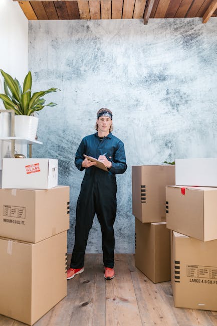 A male removal specialist from Man wth Van Rush Green is inside a residential property, holding a clipboard and noting items. He is standing among partially packed cardboard boxes, some sealed and oth