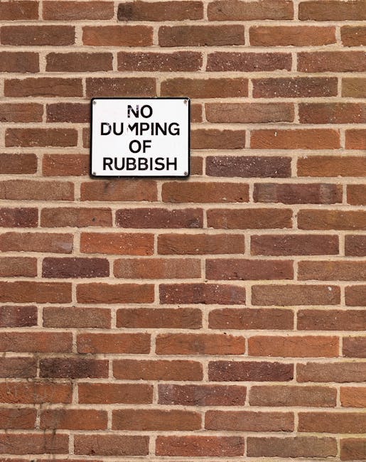 A close-up image of a red brick wall in an exterior setting, featuring a rectangular black and white sign mounted at about eye level. The sign reads 'NO DUMPING OF RUBBISH' in bold uppercase letters, with the word 'DUMPING' partially obscured due to the alignment of the sign. The wall is constructed with uniformly arranged bricks and light-colored mortar, and the lighting suggests daytime conditions with even natural light. This image depicts a typical urban or residential property boundary where waste disposal rules are enforced, relevant to house removals and waste management regulations that Man wth Van Rush Green, a house removals service, may need to adhere to when coordinating moving and disposal processes in the Barking and Dagenham area.