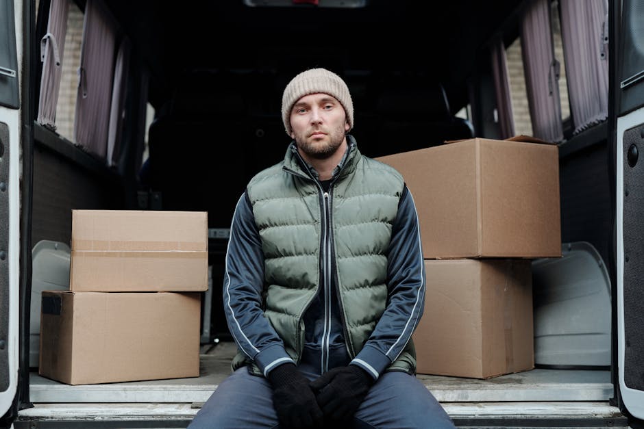 Inside the back of a white van, several cardboard moving boxes of various sizes are stacked neatly, with some boxes placed on top of others, ready for home relocation. Two individuals are present: a m