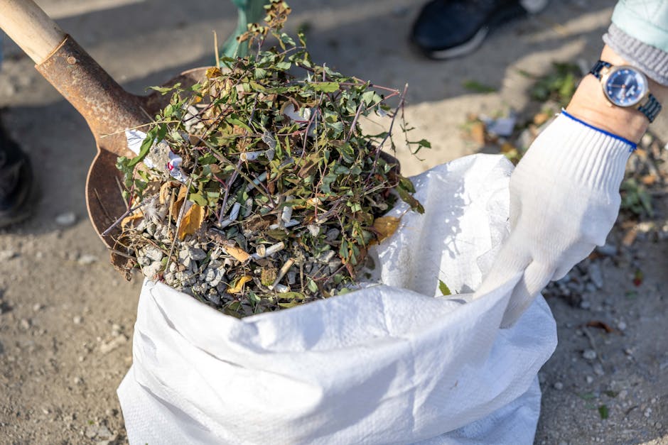 A person wearing a white glove and a watch on their wrist is holding open a large white biodegradable bag filled with garden waste, including small branches, leaves, and twigs. The bag is placed on a gravel surface, with a metal shovel positioned above it containing more garden debris. In the background, parts of men's dark shoes are visible, indicating outdoor activity related to garden or household waste collection. The scene suggests a home relocation or clearance involving the disposal of garden furniture, plants, or other waste materials as part of a removal or moving process, supported by the presence of the glove and tools likely used for packing or loading during a house move managed by Man wth Van Rush Green.