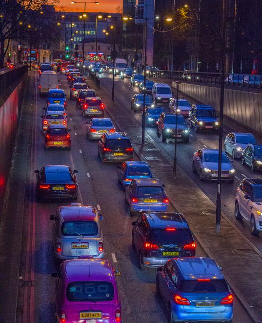 A multi-lane urban road during evening rush hour, with a high volume of stationary and slow-moving vehicles including cars, vans, and trucks, many with illuminated tail lights. The scene is visible from an elevated perspective, showing rows of cars lined up in traffic, some with visible license plates. Streetlights and traffic signals light the road, with buildings and trees in the background. On the right side, there is a sidewalk separated from the road by a guardrail, with street lamps along its length. The overall scene depicts typical urban congestion which might impact home relocation or furniture transport logistics, relevant to moving services such as those offered by Man wth Van Rush Green, especially in the context of route planning during a house move or furniture transport in a busy Rush Green area.