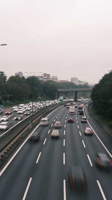 A busy multi-lane motorway with several vehicles, including cars, vans, and trucks, moving in both directions under an overcast sky. In the foreground, a black van is parked on the pavement, indicating a possible loading or unloading point related to house removals or home relocation services. Visible behind the van are cardboard boxes, some wrapped in plastic, along with additional packing materials such as blankets and plastic wraps, placed on the pavement near the vehicle. The van appears to be part of a professional moving operation by Man wth Van Rush Green, engaged in furniture transport or packing and moving activities, typical of a house removal process undergoing logistical planning and execution. The backdrop includes trees and distant buildings, with an overcast sky contributing to diffuse lighting conditions across the scene. This detailed overview captures both the urban traffic environment and the搬家相关的物品准备， reflecting typical elements involved in a situational move or furniture transport along a busy roadway. 