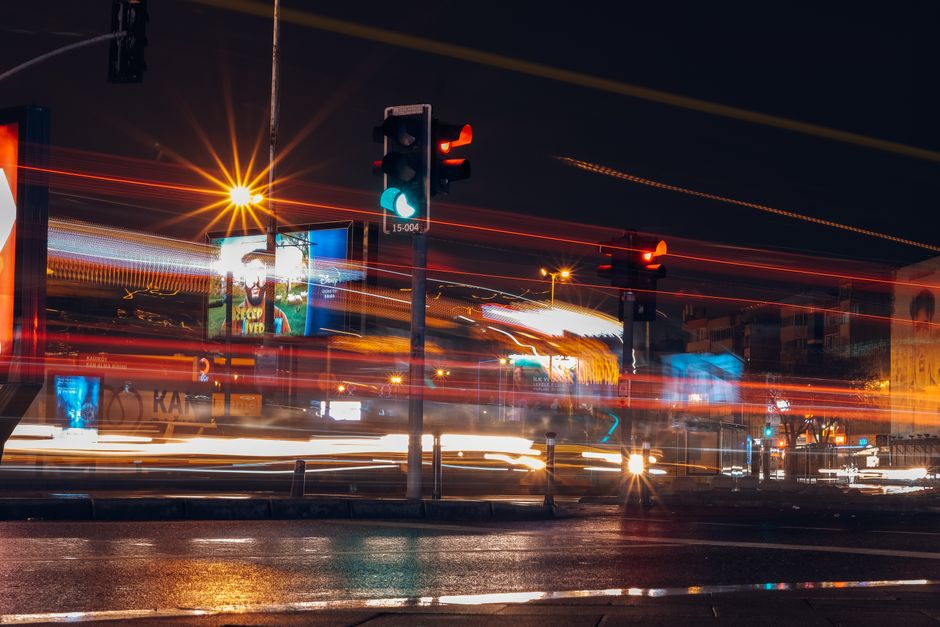 A multi-lane urban road during evening rush hour, with a high volume of stationary and slow-moving vehicles including cars, vans, and trucks, many with illuminated tail lights. The scene is visible from an elevated perspective, showing rows of cars lined up in traffic, some with visible license plates. Streetlights and traffic signals light the road, with buildings and trees in the background. On the right side, there is a sidewalk separated from the road by a guardrail, with street lamps along its length. The overall scene depicts typical urban congestion which might impact home relocation or furniture transport logistics, relevant to moving services such as those offered by Man wth Van Rush Green, especially in the context of route planning during a house move or furniture transport in a busy Rush Green area.