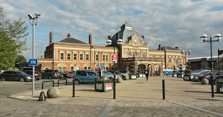 A spacious parking area in front of a historic brick building with a central clock tower and ornate architectural details, situated near Queen's Hospital Romford. The parking lot features several parked cars, including a blue van and a black sedan, along with potted plants and decorative street lamps lining the paved walkways. A blue 'IN' parking sign is visible on a pole, indicating entrance points for vehicles. The area is well-lit by natural daylight, with a partly cloudy sky overhead. In the foreground, there are bollards and large granite spheres to control vehicle access, and a trolley is partially visible near the curb. The scene depicts an organized setting suitable for home relocation activities, with ample space for loading and unloading furniture and boxes. Occasional pedestrians are seen walking, and the environment appears clean and accessible, supporting the logistics involved in a furniture transport or packing and moving process handled by Man wth Van Rush Green, providing essential parking advice for moving operations near the hospital.