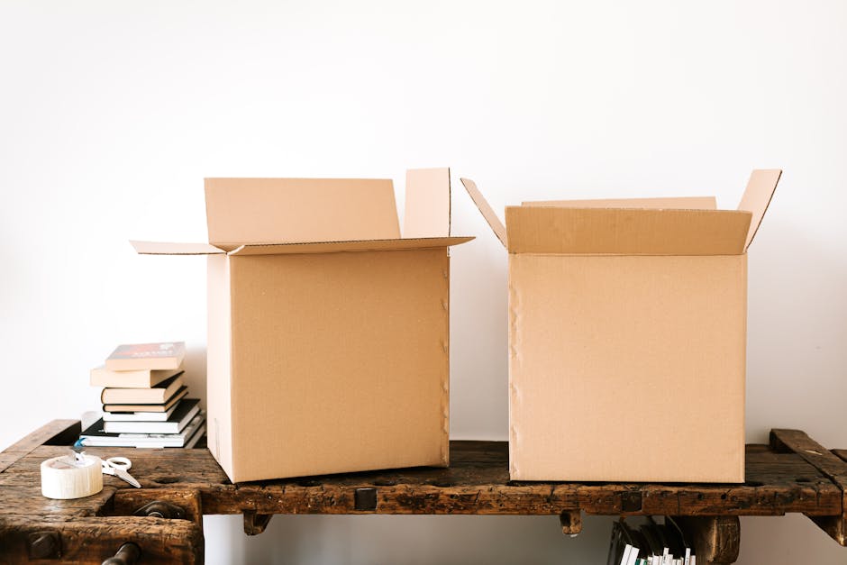 Two open cardboard moving boxes placed side by side on a rustic wooden table, with various packing materials such as white tape and scissors nearby. The box on the left contains a stack of books, whil