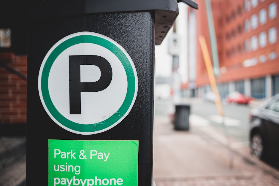 Close-up of a black parking meter with a large white circular sign displaying a black 'P' symbol, indicating parking availability. Below the sign, a green digital display reads 'Park & Pay using paybyphone,' suggesting a payment system for parking. The background shows an urban street scene with a red-brick building, parked cars, and a partially visible sidewalk. The environment is well-lit, indicating daytime, and the focus is on the parking meter, which is relevant to house removals and parking regulations in Rush Green. Man wth Van Rush Green provides removals services that often require understanding parking rules and permits for efficient furniture transport and home relocation within the area.