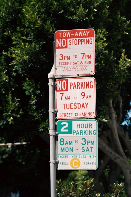 Close-up of a metal parking signpost situated outdoors in an urban environment, with three rectangular signs attached vertically. The top sign is red and white, indicating tow-away zones with no stopping allowed from 3 pm to 7 pm, except Saturdays and Sundays, with a phone number for towing inquiries. The middle sign is also red and white, restricting parking from 7 am to 9 am on Tuesdays for street cleaning. The bottom sign is green and white, indicating a two-hour parking limit from 8 am to 3 pm Monday through Saturday, with a note that vehicles with area C permits are exempt. The background features lush leafy trees and parts of a clear blue sky, providing natural lighting that highlights the signs' visibility. This signage may be relevant in the context of house removals and moving logistics managed by Man wth Van Rush Green, especially concerning parking regulations during home relocation or furniture transport activities.
