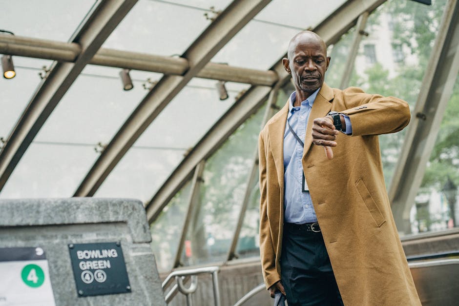 A middle-aged man in a light brown overcoat and blue shirt stands outdoors inside a modern glass-covered area, likely a transit station or part of a home entrance. He is checking his smartwatch with his right hand while holding a black bag or briefcase in his left hand, appearing to be in a waiting or checking process. Behind him, the curved glass roof structure and metal supports are visible, allowing natural daylight to illuminate the scene. The background features blurred greenery and a paved surface, suggesting an urban environment with accessible outdoor space. This scene is relevant to home relocation or moving services, illustrating the logistical aspect of timing and planning involved in house removals. Such a setting may be part of client coordination, travel planning, or the transportation process managed by companies like Man wth Van Rush Green, highlighting the importance of punctuality and organized logistics in furniture transport or packing and moving operations.